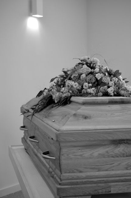 A polished wooden casket with a bouquet of fresh flowers resting on top, placed on a stand against a plain wall illuminated by a small wall-mounted light. The floral arrangement includes light-colored