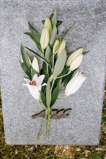 A floral tribute featuring white lilies and white enchanters, arranged with green leaves and stems, affixed to a textured gray stone surface. The lilies are in various stages of bloom, with some petal