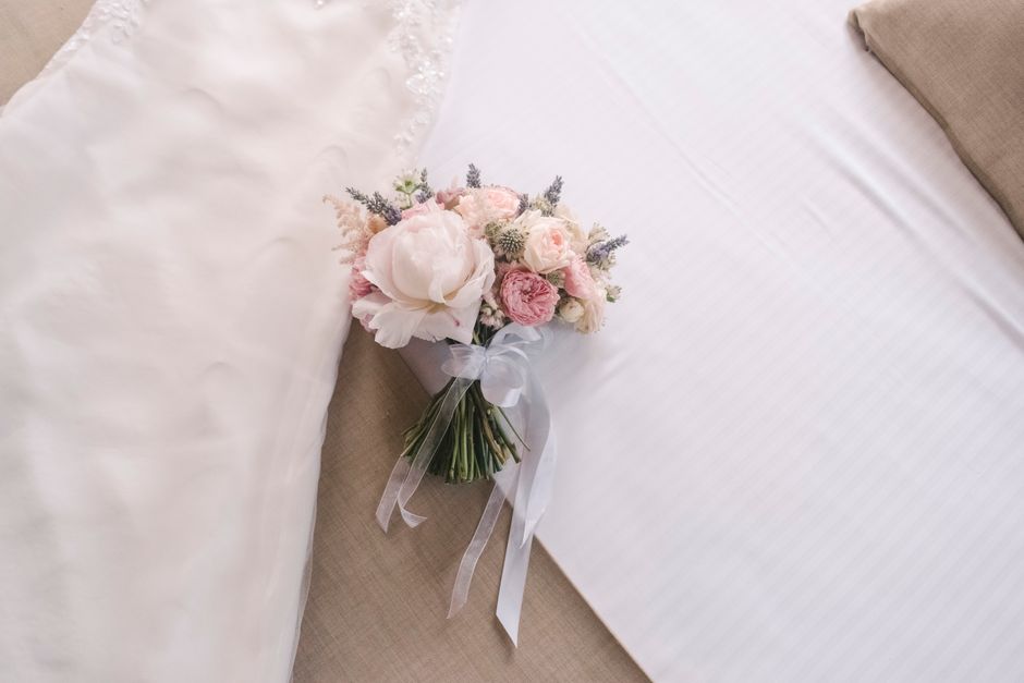 A delicate bouquet of fresh flowers, including soft pink roses, white peonies, and lavender sprigs, arranged with greenery. The bouquet is wrapped in white tissue paper, tied with a sheer white ribbon