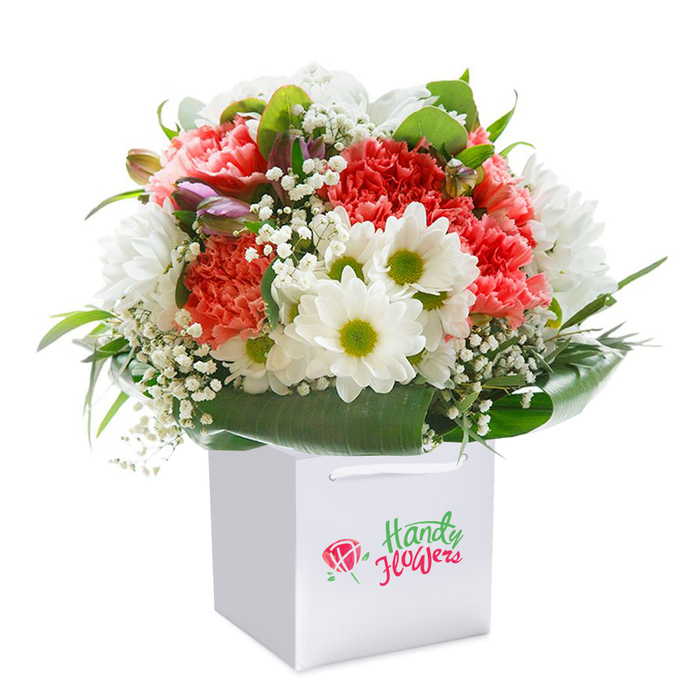 This striking arrangement, nestled upright in a crisp white square gift bag, exemplifies the distinct floral artistry found in Wealdstone. At the heart of the bouquet lie lush coral and peach carnations whose ruffled, full petals impart a welcoming warmth, offset by a scattering of snowy white daisies, each displaying a vibrant lime-green core. Velvet petals and crisp textures blend seamlessly as delicate sprays of baby's breath float among the primary blooms, adding both softness and a light, gentle fragrance evocative of a stroll through the gardens off Kenton Road. Tucked subtly to the left, you'll spot a few purple-tinged buds-just hinting at the next chapter of spring, while the arrangement's lush collar of deep green, glossy leaves provides supportive structure and rich contrast. The white gift bag, with its ribbon handles, stands as a modern counterpoint to the natural shapes inside and is perfect for safe, elegant transport-whether destined for a gathering near Wealdstone's Clock Tower or a quiet get-together at home. Presented against a pure white backdrop, every aspect of this bouquet radiates freshness, balance, and thoughtful artistry typical of the Wealdstone area, making it a fitting centerpiece or cherished gift for celebrations, birthdays, or simple everyday joys.