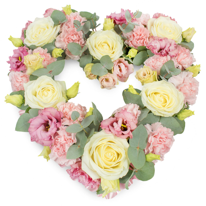 Against a bright, uncluttered white backdrop sits a full, heart-shaped wreath created from a harmonious blend of pastel flowers and cool greenery. The heart is symmetrical and clearly defined, every inch of its outline filled with blossoms that touch edge to edge. Large cream and pale lemon roses serve as anchors within the design, their spiralled petals forming calm, rounded centres that immediately catch the eye. Weaving around these roses are generous clusters of light pink carnations, their ruffled petals layered in soft profusion, adding a sense of volume and tenderness to the heart. Punctuating the pastel scene are deeper pink and mauve lisianthus, their more open, gently fluted petals providing contrast in both shape and tone, like small echoes of evening light. Slender stems of eucalyptus thread through the arrangement, the silvery-green, slightly rounded leaves peeping through the flowers with a cool, soothing presence. Here and there, tiny yellow-green buds stand upright, suggesting new life and freshly cut stems chosen with care. The overall effect is lush yet gentle, as if the flowers have been lovingly placed one by one. This is the kind of tribute families around Wealdstone might choose for a church service, a crematorium farewell, or a quiet remembrance at home, when they want something that conveys affection, respect, and enduring connection. The white background emphasises the softness of the pinks and creams, allowing the heart shape-and the sentiment behind it-to take centre stage without distraction.